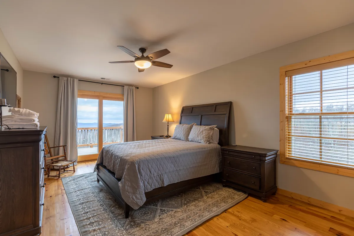 A plush bed dressed in soft gray bedding sits beside a large window offering expansive, beautiful views of the outdoors. The wooden furniture and gentle lighting create a cozy, serene atmosphere—ideal for unwinding after a day of exploration or for evening stargazing from the comfort of the suite.