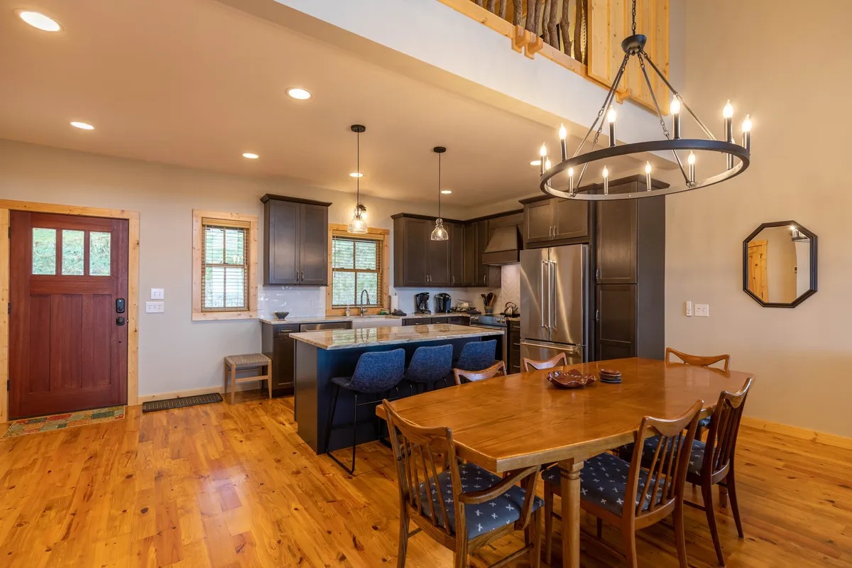 The scene centers on a spacious dining table surrounded by comfortable chairs, with an island counter and attractive kitchen cabinetry nearby. Warm light from a chandelier and sunlight streaming in add to the family-friendly, communal dining mood.