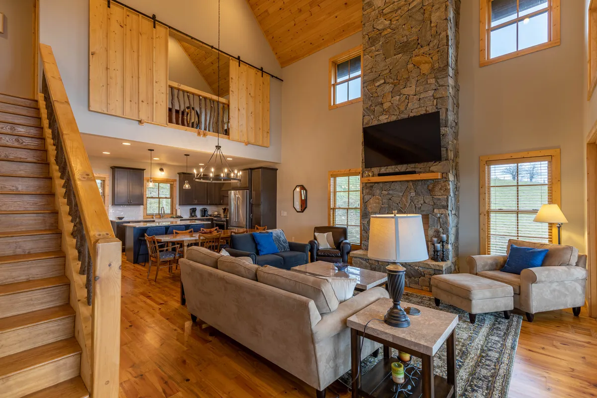 A wider view of the living area shows the stone fireplace, open staircase, comfortable sofas, and adjacent dining space. The room exudes warmth with tongue-and-groove woodwork and vaulted ceilings, capturing the open-concept design and the flow between gathering spaces.