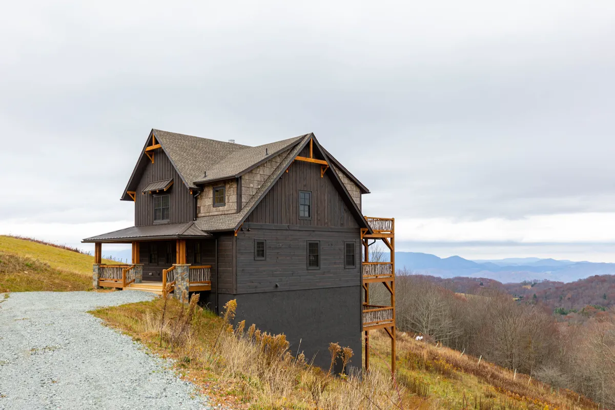 A broad view of the home's exterior, with a tranquil mountain backdrop and a driveway leading up to the house. The image emphasizes the connection to nature, with open sky, greenery, and the welcoming presence of the house nestled in its setting.
