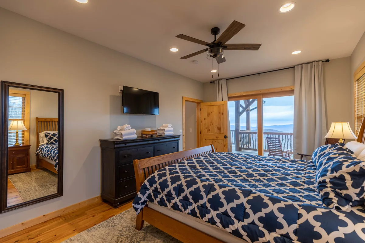A serene bedroom featuring a comfortable bed with a patterned duvet, a dresser with neatly arranged towels, a generous mirror reflecting the space, and a sliding glass door leading to a private balcony. The room radiates calm, with opportunities for relaxation, enjoying the view, or starting the day with fresh air.