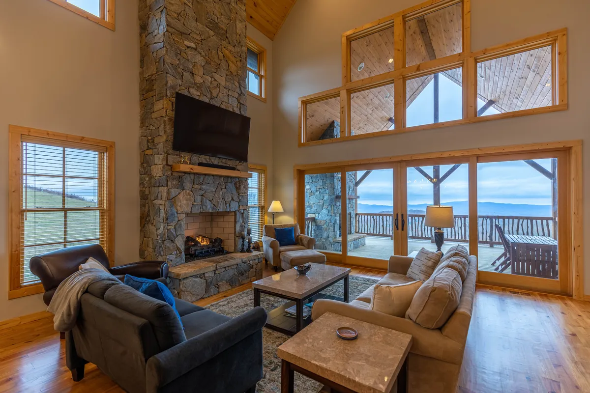 This image captures the great room's stone fireplace as a central focal point, flanked by large windows bathing the space in natural light. The comfortable seating area features two plush sofas, inviting guests to gather for relaxation. Through the windows, expansive views of valleys and peaks are visible, emphasizing the connection to the outdoors.