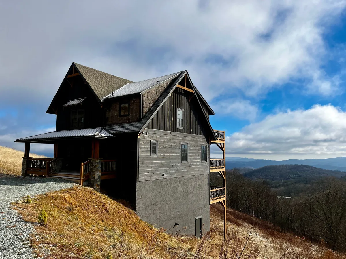 A rustic wooden balcony with dark siding and a stone foundation overlooks a stunning mountain panorama. The scene evokes feelings of peacefulness and connection to nature, ideal for enjoying sunrise or sunset moments.