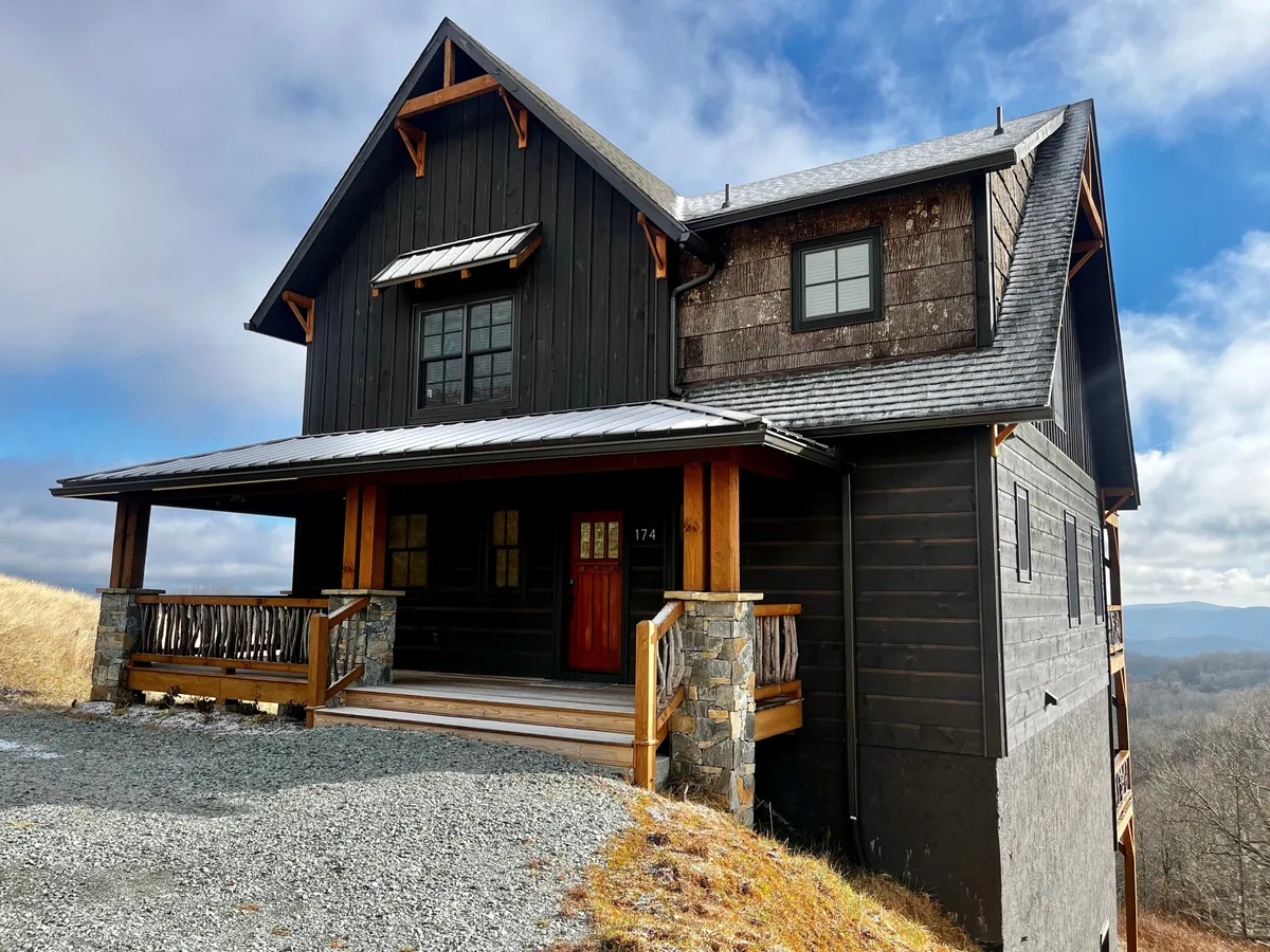 The image showcases the home's inviting wooden porch with stone foundation and dark wood siding, framed by large windows that reveal expansive mountain views. The scene exudes warmth and comfort, perfect for relaxing outdoors, stargazing, or gathering with loved ones while surrounded by the beauty of the North Carolina mountains.