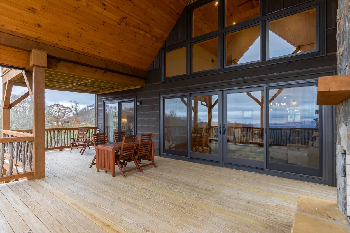 A welcoming wooden deck featuring comfortable outdoor seating and rustic railings, with large glass doors connecting to the indoor living area. The deck is partially covered, providing a cozy space for gathering with friends or relaxing while enjoying the surrounding views.