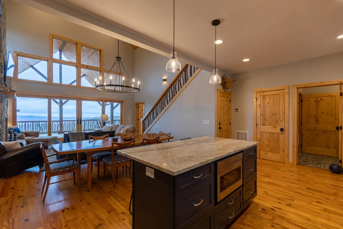 A wide view that includes both the dining table and kitchen island, with large windows that invite natural light. The scene suggests a seamless connection between cooking, dining, and gathering, with the staircase hinting at the home's open flow.